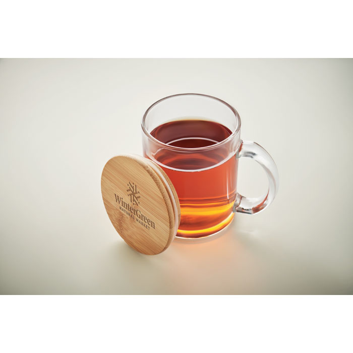 A glass transparent mug with a bamboo lid (light brown), filled with tea, and the lid is engraved with a logo 